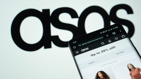 Photo of a smartphone open to the Asos website. The retailer's logo is displayed in black on a slightly off-focus white backdrop behind the phone.